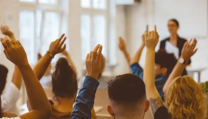 Schüler heben die Hände, um Fragen zu stellen. Ein achtsamkeit lehrer im Hintergrund beobachtet die Klasse.