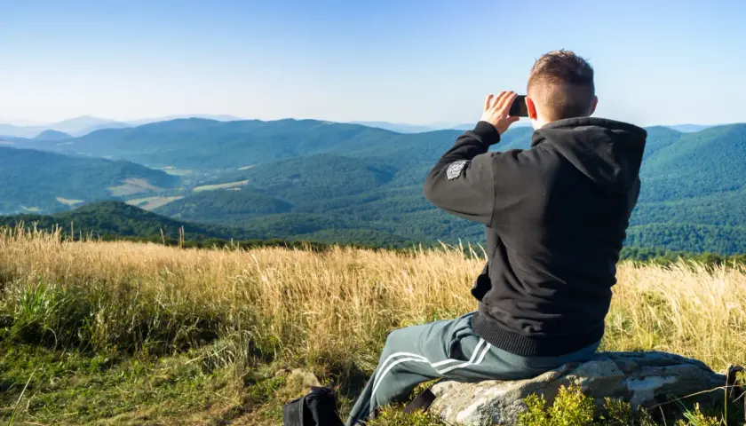 Mężczyzna siedzi na skale, robiąc zdjęcie krajobrazu telefonem z aparatem szerokokątnym.