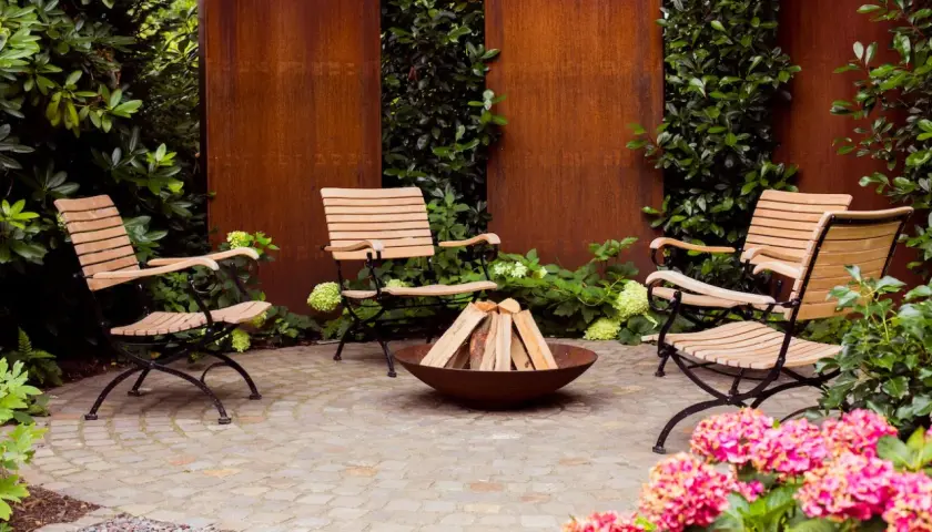 Moderne, schlichte Gartengestaltung mit vier Holzstühlen um eine Feuerstelle. Rostige Metallwände und üppiges Grün schaffen eine gemütliche Atmosphäre.