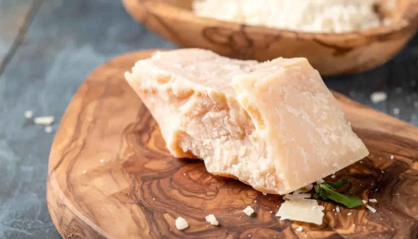 Kawałek twardego sera, przypominającego Grana Padano i parmezan, leży na drewnianej desce. W tle widać misę z tartym serem.