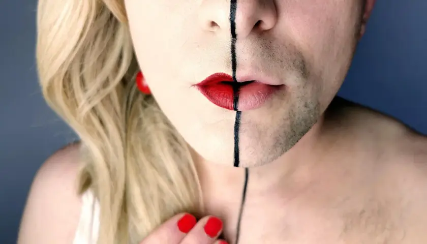 Un volto diviso a metà, con capelli biondi e barba, labbra rosse e una linea nera. Un'espressione di una persona non binaria che abbraccia la propria identità.