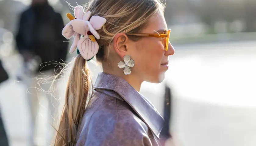 Mujer con coleta adornada con una flor de tela y pendientes de pétalos, luciendo gafas naranjas y una chaqueta de cuero.