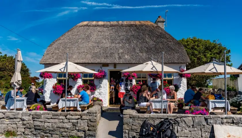 Café pittoresque avec toit de chaume, parasols et fleurs. Des gens dégustent un repas en plein air, peut-être un délicieux aran poisson.