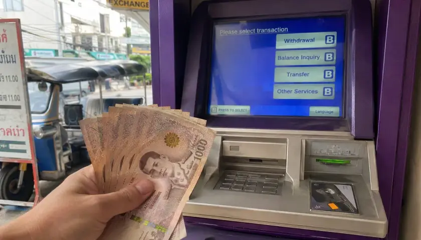 Hand hält thailändische Baht-Scheine vor einem Geldautomaten. Hier kann man Geld wechseln.