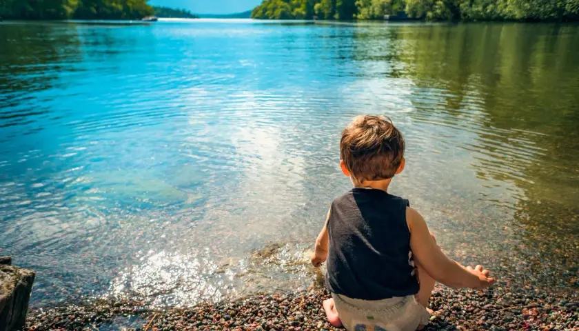 Un bambino seduto sulla riva di un lago, godendosi un tranquillo week end con bambini. L'acqua limpida riflette il cielo azzurro e gli alberi verdi.