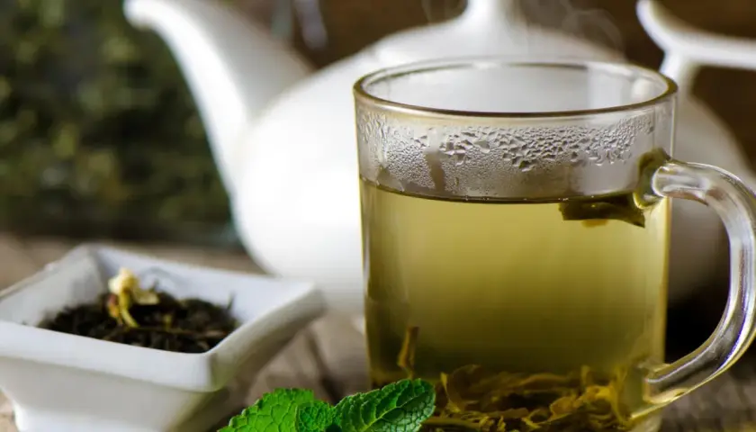 Dampfender grüner Tee in einer Tasse, daneben eine Schale mit Teeblättern und Minze. Welcher Tee ist antibakteriell? Grüner Tee ist bekannt für seine gesundheitlichen Vorteile.