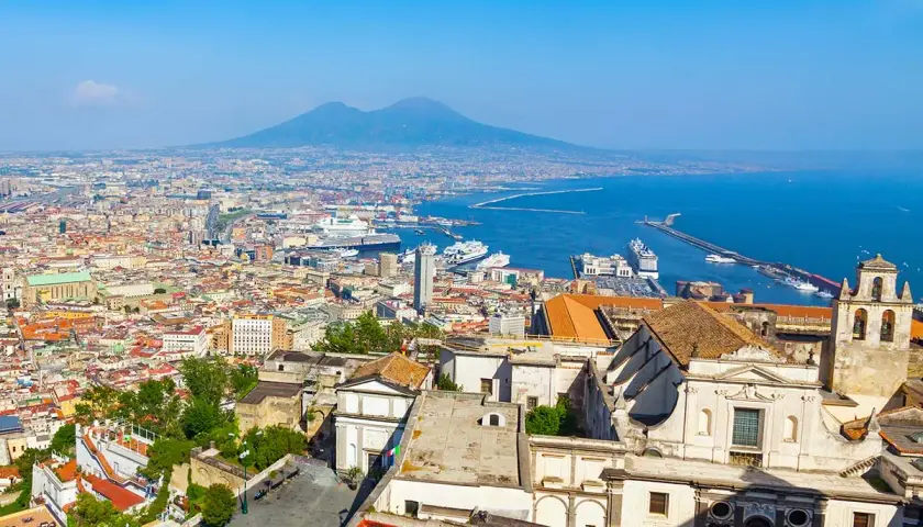 One day in Naples, a panoramic view reveals the city's sprawl, the bustling port with cruise ships, and the majestic Mount Vesuvius in the distance under a clear blue sky.