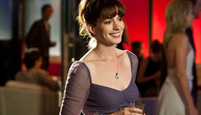 Uśmiechnięta Anne Hathaway w fioletowej sukience, trzymająca drinka. Scena z jednego z jej filmów.