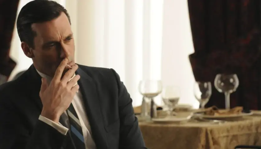 Jon Hamm, znany z filmów, seriali i programów, w eleganckim garniturze pali papierosa przy stole.