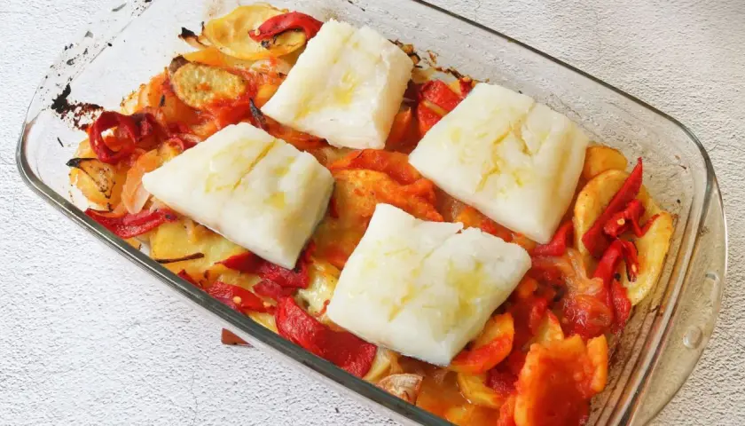 Cuatro porciones de bacalao al horno reposan sobre patatas y pimientos asados en una fuente de vidrio.