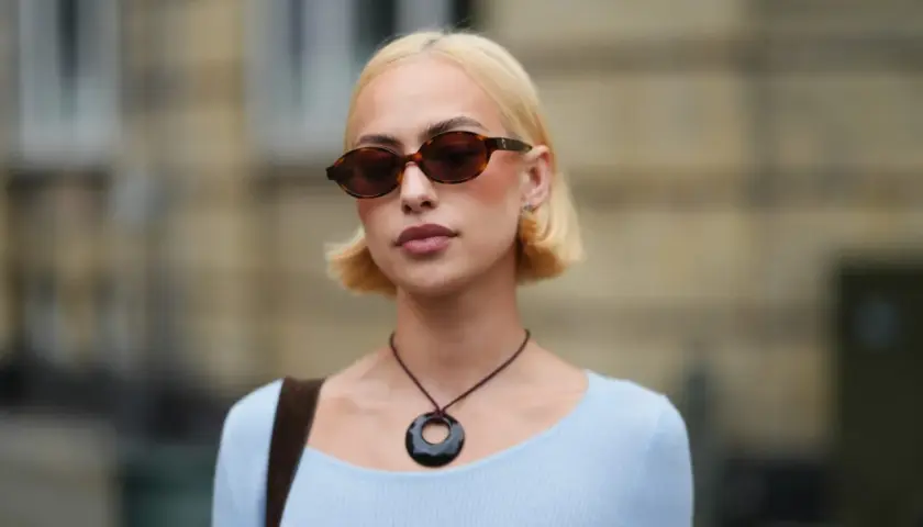 Krótkie fryzury bob w blondzie, stylowe okulary i naszyjnik z okrągłą zawieszką.