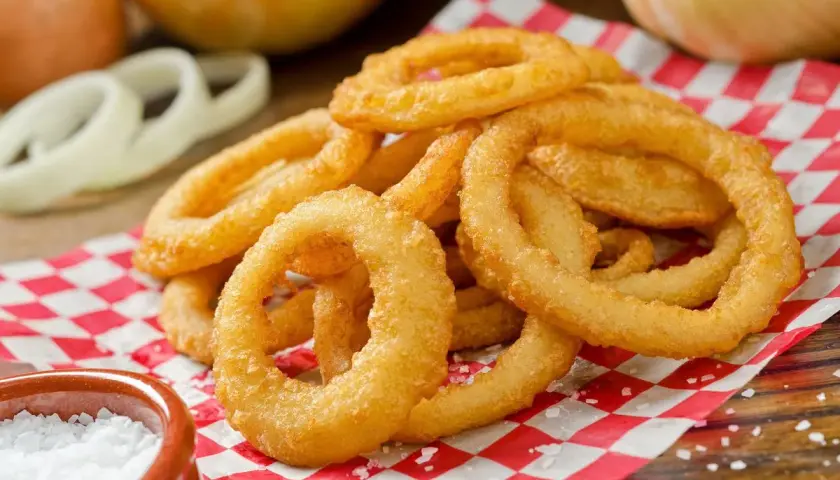 Anelli di cipolla dorati e croccanti, preparati con pastella con acqua frizzante, serviti su carta a quadri rossi e bianchi, con sale grosso a lato.