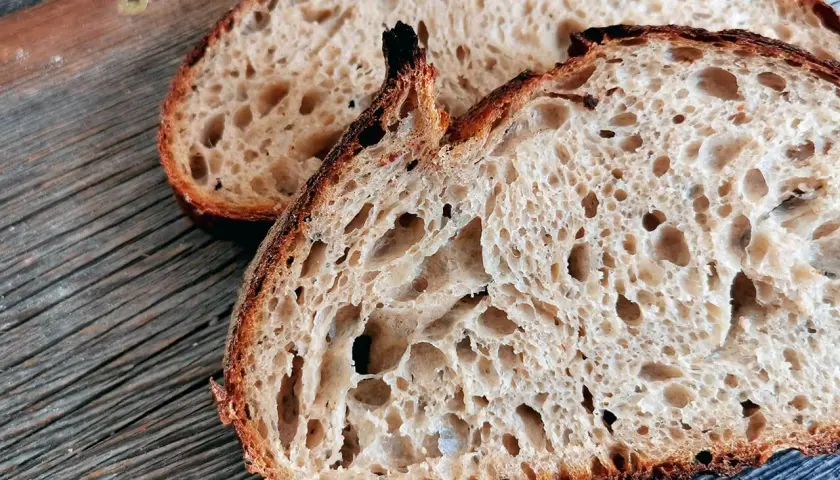 Zwei Scheiben rustikales Sauerteigbrot mit großer Porung liegen auf einem Holzbrett. Warum ist Sauerteig gesund? Weil die Fermentation Nährstoffe besser verfügbar macht.
