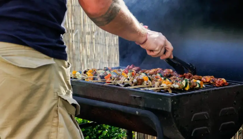 Un homme utilise des pinces pour retourner des brochettes colorées sur un barbecue fumant. C'est le moment idéal pour degraisser le barbecue après ce festin !