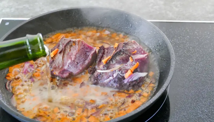 Deux morceaux de joue de bœuf mijotent dans une poêle avec des carottes et des oignons. Un liquide est versé d'une bouteille verte.