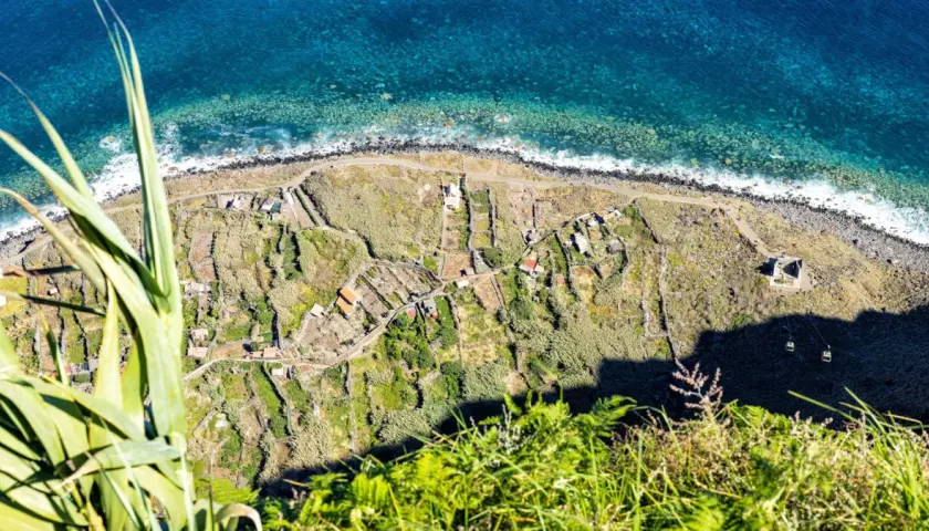 Terraced fields and houses dot the coastline, a glimpse of what to see in Madeira. The turquoise ocean crashes against the rocky shore.