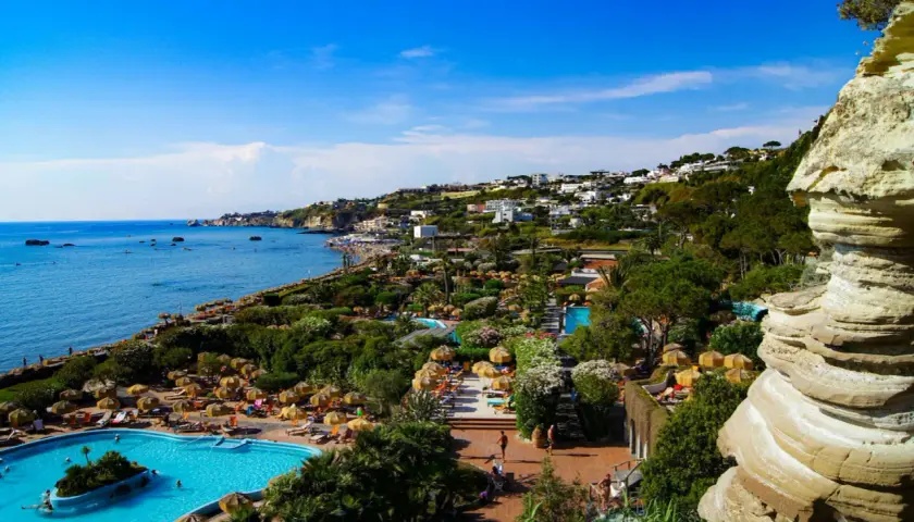 Vista panoramica dell'isola d'Ischia con mare azzurro, piscine, vegetazione lussureggiante e case arroccate sulla collina.