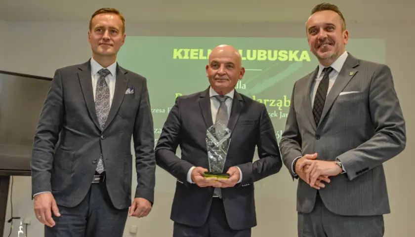 Trzech mężczyzn w garniturach pozuje do zdjęcia, jeden z nich trzyma statuetkę.