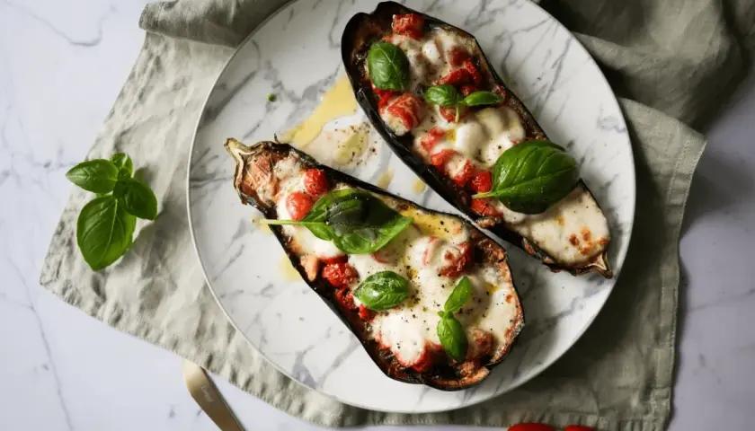 Dwa połówki bakłażana pieczonego z pomidorkami i mozzarellą, udekorowane listkami bazylii.