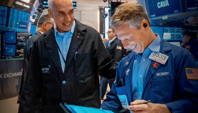 Dwóch mężczyzn w kurtkach NYSE śmieje się, patrząc na tablet. Jeden z nich, Bobby, pokazuje coś na ekranie, a drugi, z napisem "skup akcji własnych" na piersi, uśmiecha się szeroko.