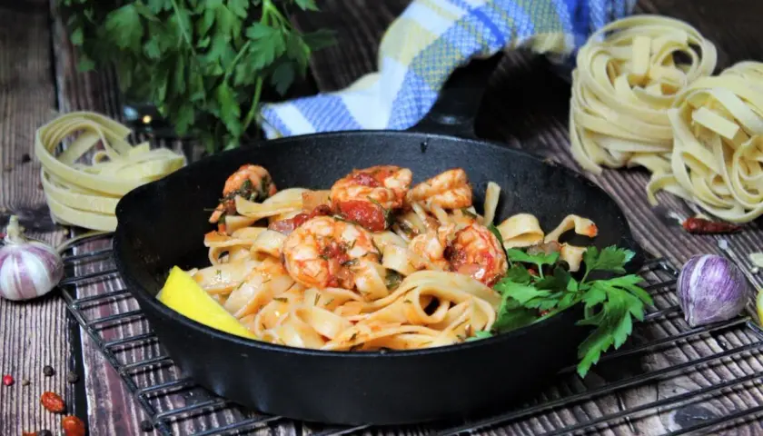 Spaghetti mit Garnelen mediterran in einer Gusseisenpfanne, garniert mit Zitrone und Petersilie. Daneben rohe Pasta, Knoblauch und Chili.