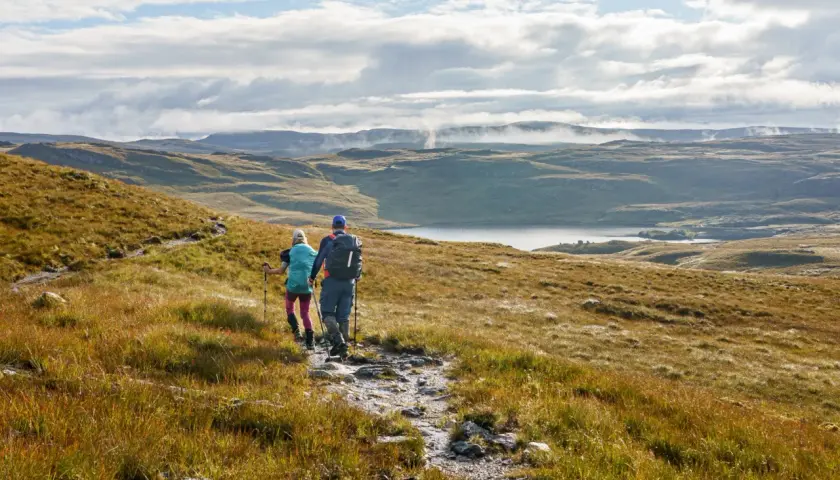 Two hikers trek along the Cape Wrath Trail, a rugged path winding through a vast, misty Scottish landscape with a serene loch in the distance.