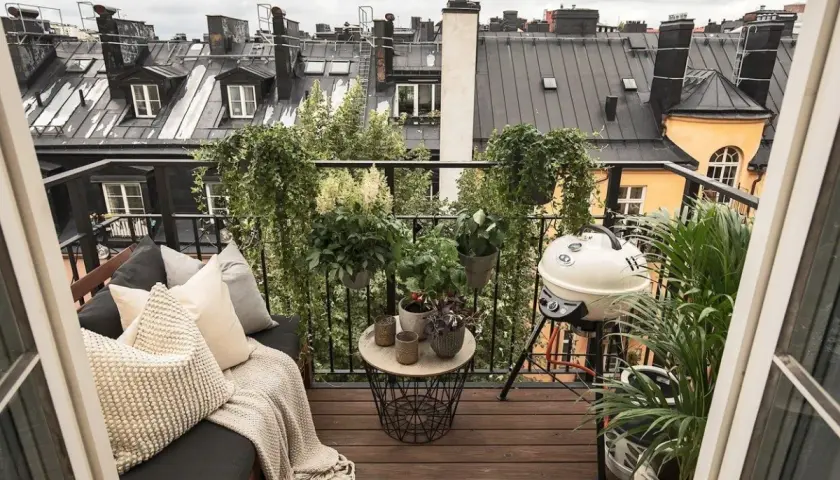 Aménagement terrasse appartement : un coin salon confortable avec coussins, une table basse, un barbecue et de nombreuses plantes, offrant une vue sur les toits.