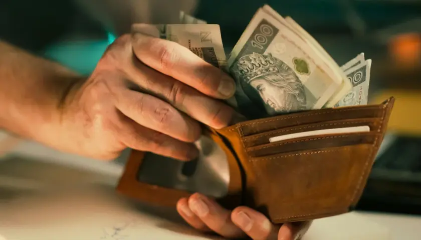 Dłonie wkładają polskie banknoty do brązowego portfela. Widać fragmenty banknotów 100 zł. Czy to już cała pensja, czy tylko minimalna brutto?