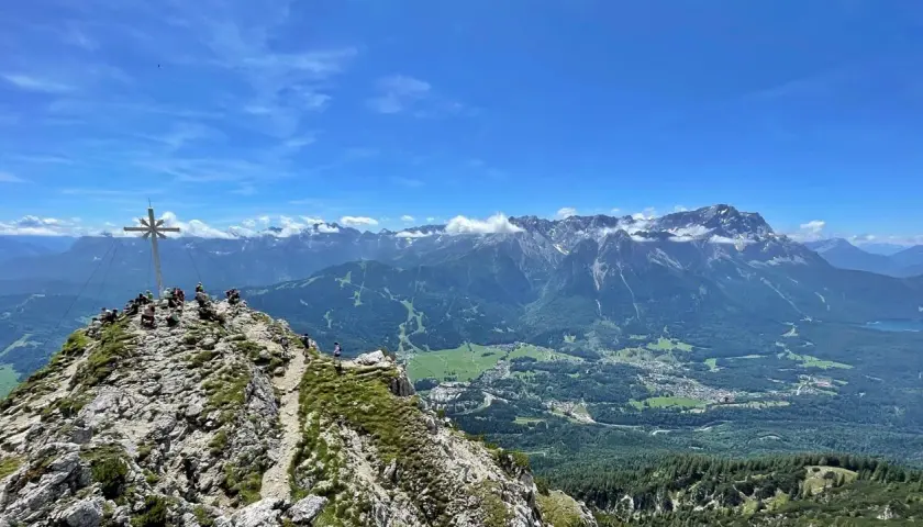 Gipfelkreuz auf dem Kramer­spitz, umgeben von Wanderern. Im Hintergrund das Alpenpanorama und ein Tal mit Häusern.
