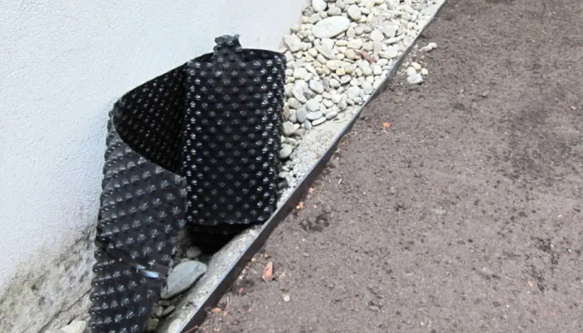 Schwarze Noppenbahn schützt die Hauswand vor Feuchtigkeit, daneben Kies und Erde.