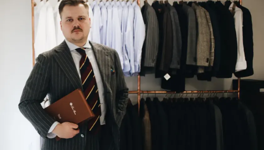 Elegancki mężczyzna w garniturze w prążki trzyma teczkę. Za nim wiszą marynarki i koszule.
