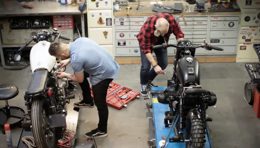 Due uomini lavorano su due moto in un garage. Uno è chino su una moto bianca, l'altro su una nera. Un kit di attrezzi è aperto sul pavimento. Progetto schienalino moto fai da te.
