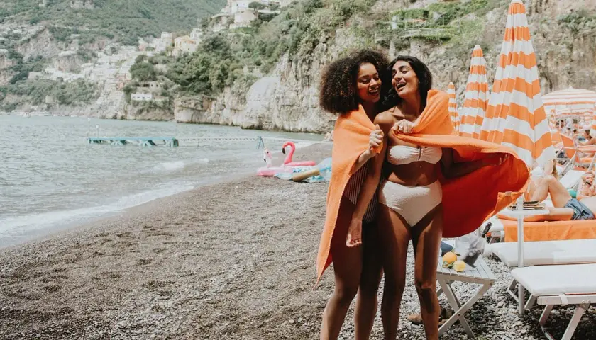 Deux amies rient sur une plage italienne, partageant une serviette orange. Leurs vacances en Italie en famille au bord de la mer sont idylliques.