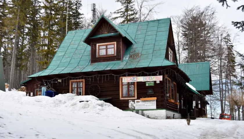 Drewniany domek z zielonym dachem, otoczony śniegiem i lasem. Na ścianie wisi baner "BĘDZIE DOBRZE", a przed wejściem stoi kot. Taki widok to prawdziwy przegibek!
