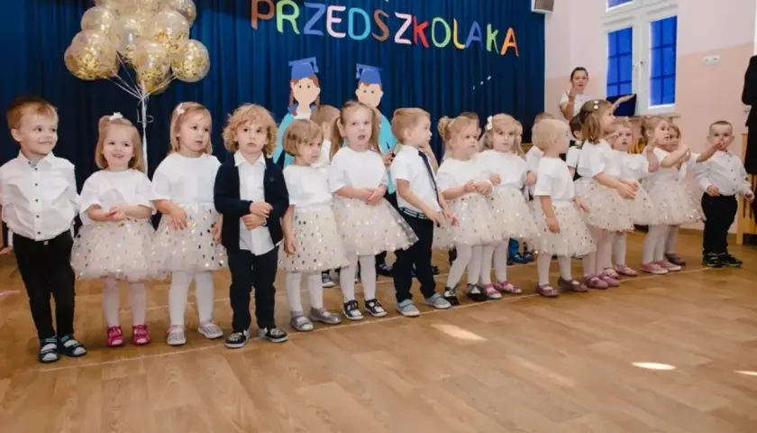 Dzieci w strojach galowych stoją w rzędzie na tle niebieskiej kurtyny z napisem "Pasowanie na przedszkolaka".
