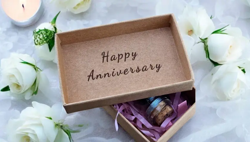 Otwarta szkatułka z napisem "Happy Anniversary" i małą buteleczką w środku, otoczona białymi różami i płonącą świecą. Idealny prezent na 21 rocznicę ślubu.