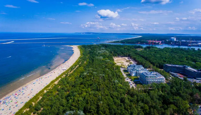 Plaża w Świnoujściu z lotu ptaka. Tłumy plażowiczów, turkusowe morze i zielony las otaczający nowoczesne apartamenty.