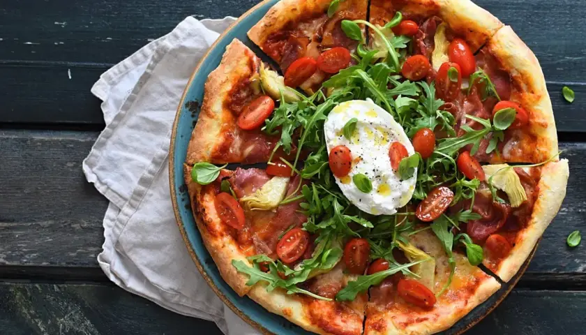 Pizza z mortadelą, rukolą, pomidorkami i burratą na niebieskim talerzu.