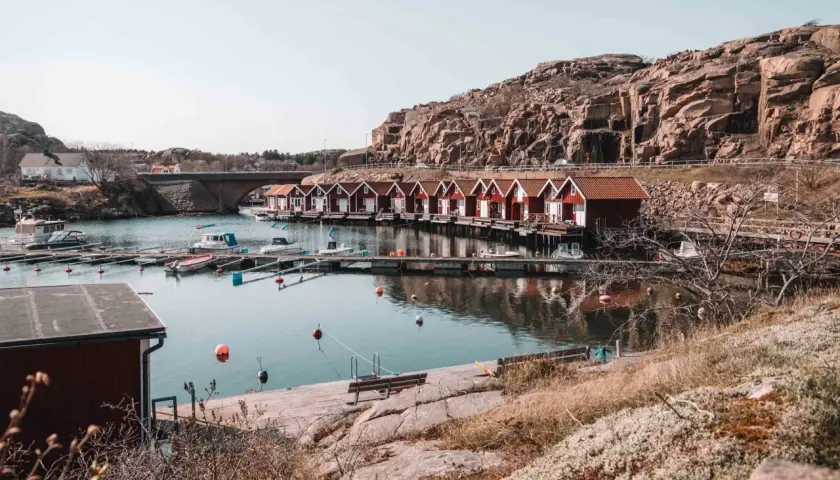 En rad röda sjöbodar längs en kuststig, med båtar i vattnet och en klippig bakgrund.