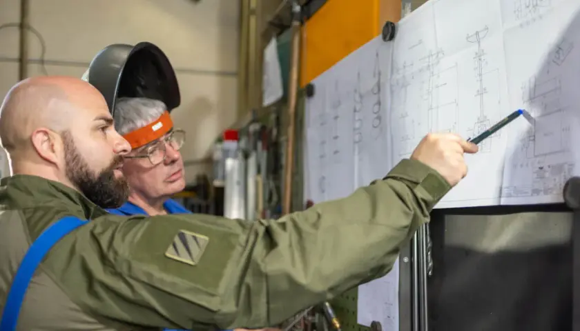 Zwei Männer besprechen einen technischen Plan. Einer zeigt mit einem Stift auf Details, die an einen **Ratschenzug** erinnern.