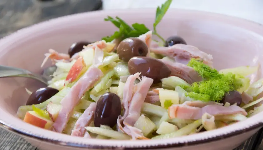 Frischer Fenchel Salat mit Apfelstücken, Schinkenstreifen und dunklen Oliven in einer rosa Schüssel.