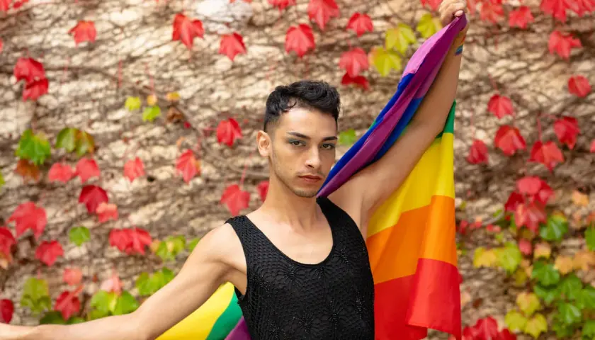 Un giovane con la bandiera arcobaleno, simbolo del **queer significato** di libertà e identità, si staglia contro un muro fiorito.