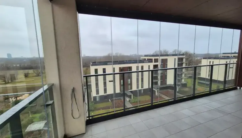 Zabudowa balkonu w bloku z widokiem na nowoczesne budynki. Szklane panele tworzą przestronną loggię.