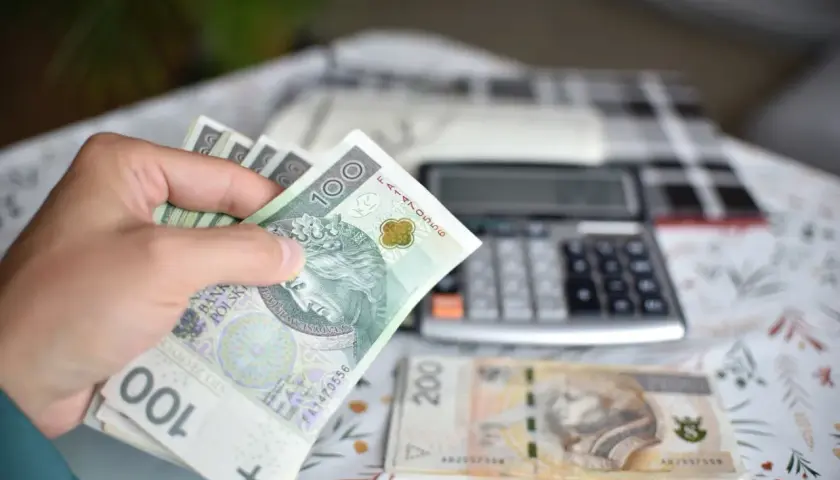Ręka trzyma banknoty 100 zł, obok leży 200 zł i kalkulator. Obliczenia ekwiwalentu za urlop.