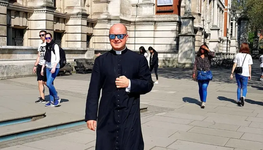 Uśmiechnięty pastor w czarnej sutannie i okularach przeciwsłonecznych idzie ulicą. W tle widać ludzi i budynek.