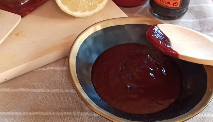 Cuchara de madera prueba la deliciosa salsa barbacoa casera en un cuenco. Limón y botellas de ingredientes al fondo.
