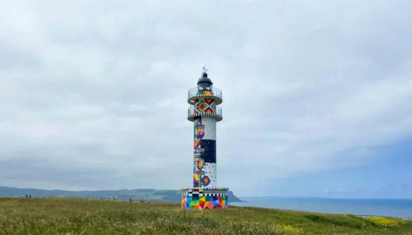 Faro colorido en el cabo de Ajo, con un paisaje verde y el mar al fondo.