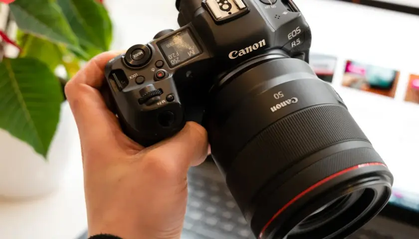 Dłoń trzyma aparat Canon EOS R5 z obiektywem 50mm. W tle widać ekran laptopa.
