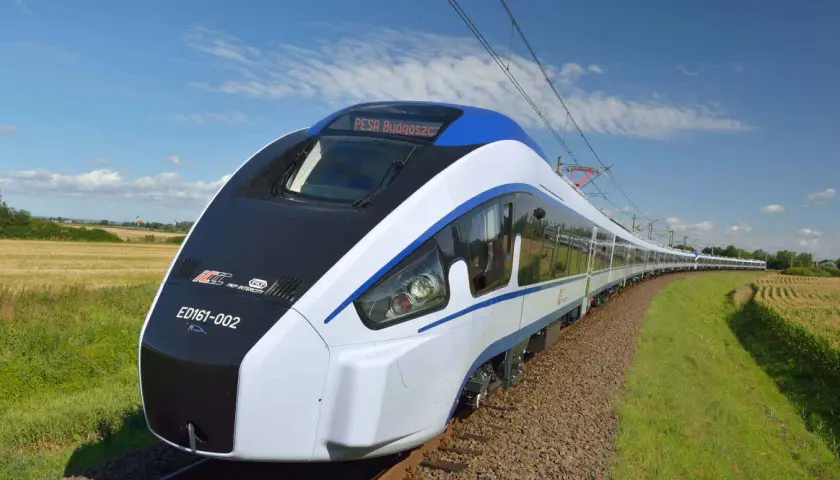 Nowoczesny pociąg Pesa Bydgoszcz ED161-002 mknie po torach wśród pól i zieleni, pod błękitnym niebem z chmurami.