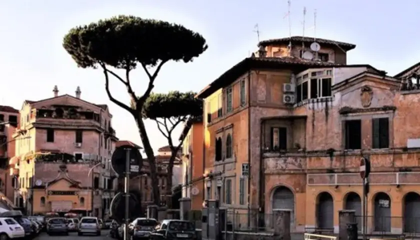 Edifici color ocra e rosa con pini marittimi svettanti, tipici de **la Garbatella**, Roma. Auto parcheggiate lungo la strada.